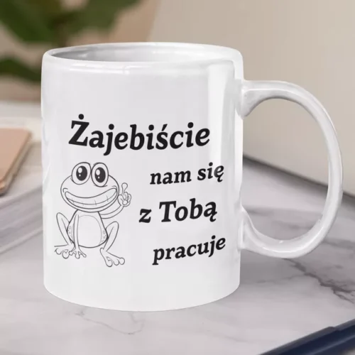 Biały ceramiczny kubek z napisem Zajebiście nam się z Tobą pracuje oraz ilustracją uśmiechniętej żaby na marmurowym blacie.