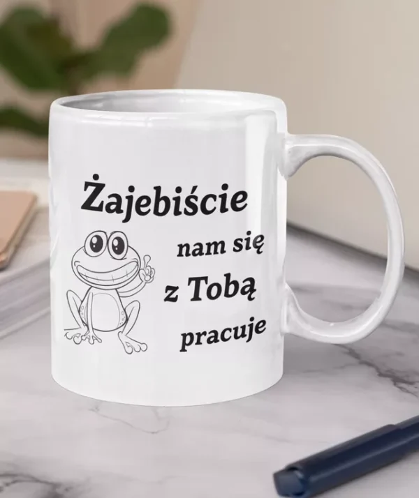 Biały ceramiczny kubek z napisem Zajebiście nam się z Tobą pracuje oraz ilustracją uśmiechniętej żaby na marmurowym blacie.